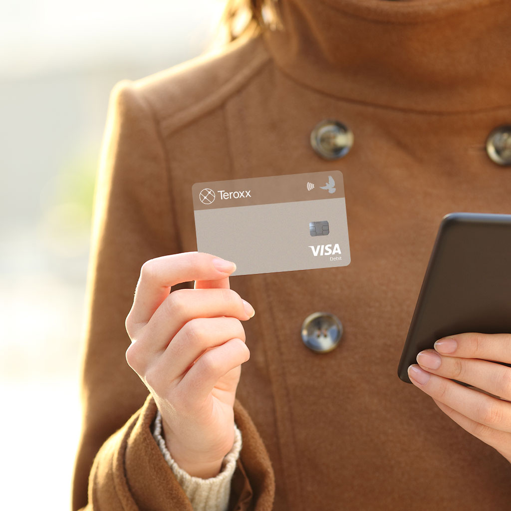 Person wearing a brown coat holding a smartphone in one hand and a transparent Teroxx Visa card in the other, outdoors.