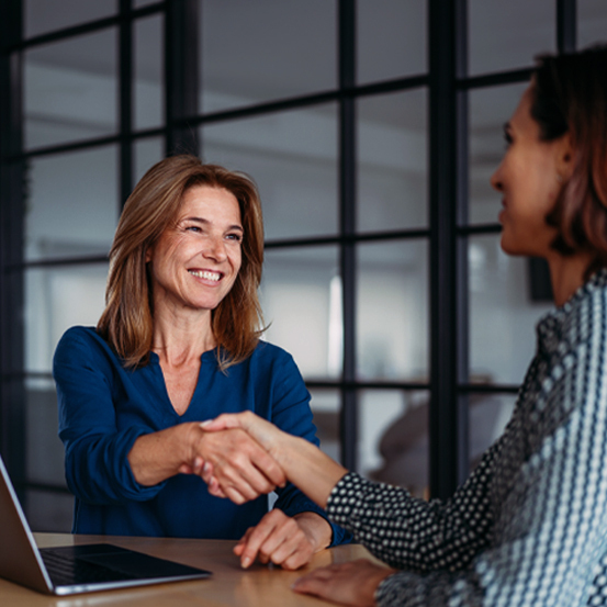 Two women in an office shaking hands across a desk, smiling during a business meeting