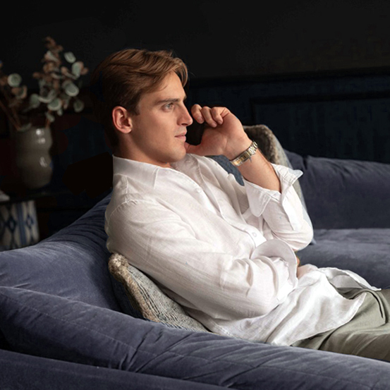 Man sitting on a velvet sofa, wearing a white shirt and watch, talking on the phone in a relaxed setting.