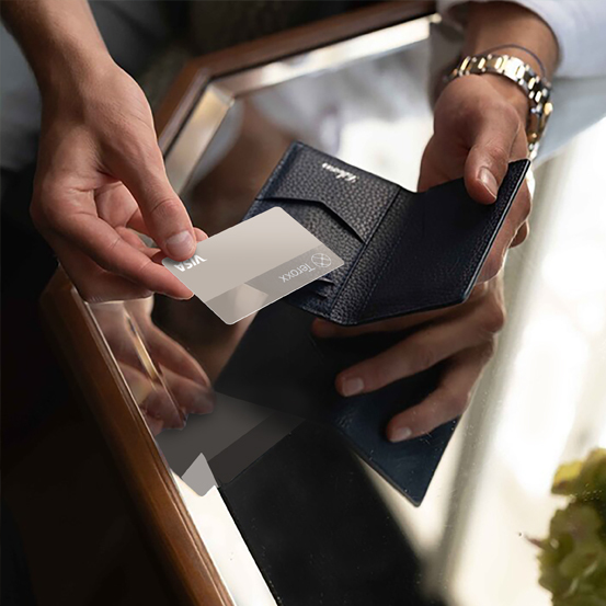 A person holding a dark leather wallet while inserting a silver Teroxx Visa card, with the reflection visible on a glass table.