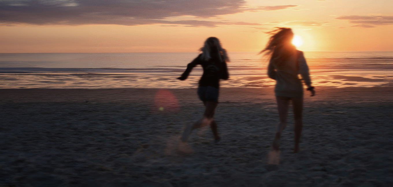 Two women run on a sandy beach towards the ocean as the sun sets in a blur of motion. Two women run on a sandy beach towards the ocean as the sun sets in a blur of motion.