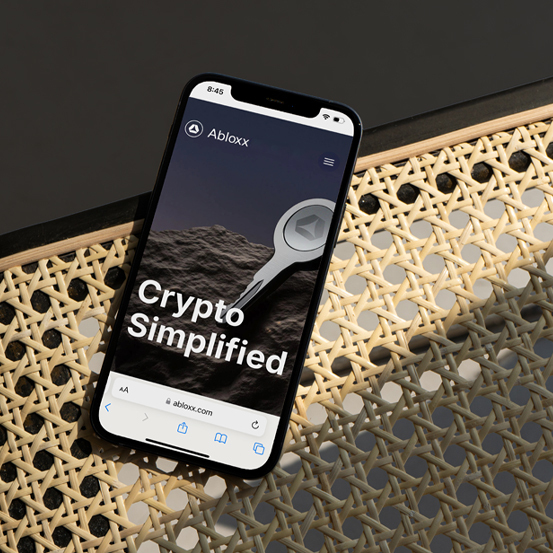 A smartphone rests on a wicker chair, displaying the Abloxx website with the text "Crypto Simplified" on the screen.