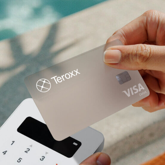 A hand holding a sleek Teroxx Visa Debit card near a card reader, with a soft background featuring sunlight and poolside vibes.