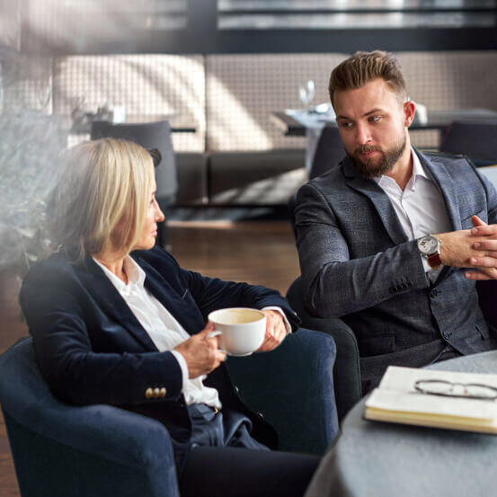 Two colleagues in business attire engage in a conversation at a modern café. One holds a coffee cup. A notebook and pair of glasses rest on the table.