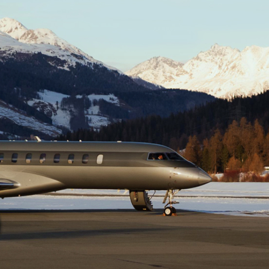 A private jet sits on an airport tarmac with a backdrop of snow-covered mountains and pine trees.