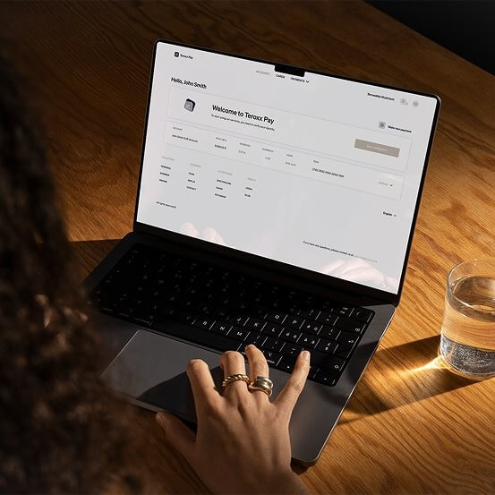 Person using a laptop on a wooden table, viewing the Teroxx Pay dashboard with a welcome message and account details visible on the screen; a glass of water is placed nearby.